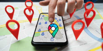An image of a person's hand pointing at a smartphone. The screen displays a map with various red location pins indicating different places of interest or destinations. The map seems to be from a digital mapping service, enhanced with colorful icons, suggesting the use of AI for travel features. The background is a larger map with several red pins, and it appears to be a physical printout, contrasting with the digital version on the phone. Google enhances Maps with new AI travel features