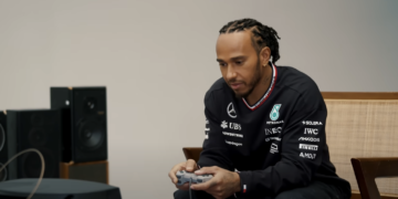 Lewis Hamilton is sitting down playing a PS1 console on a small TV screen.